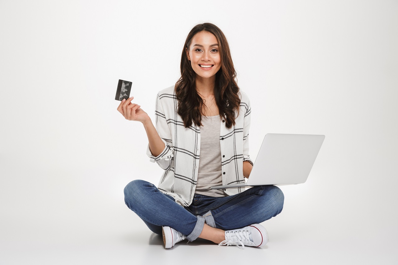 Smiling brunette woman in shirt sitting on the floor with laptop computer and credit card